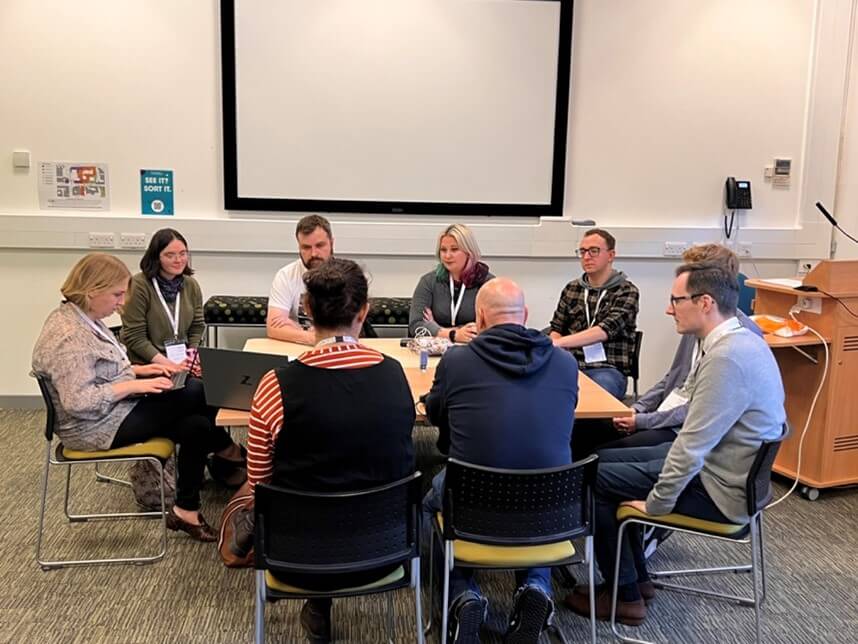 The image shows a group of delegates in a break out session gathered around a table listening to audio from a laptop.